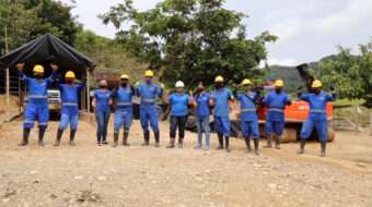 Group of men and women gold miners in blue uniforms and yellow helmets standing in front of mining equipment in a mining area on a rocky shore in a rainforest in Antioquía, Colombia.