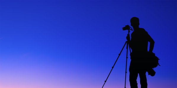 Silhouette of a person holding a tripod with a camera on it against a blue background.