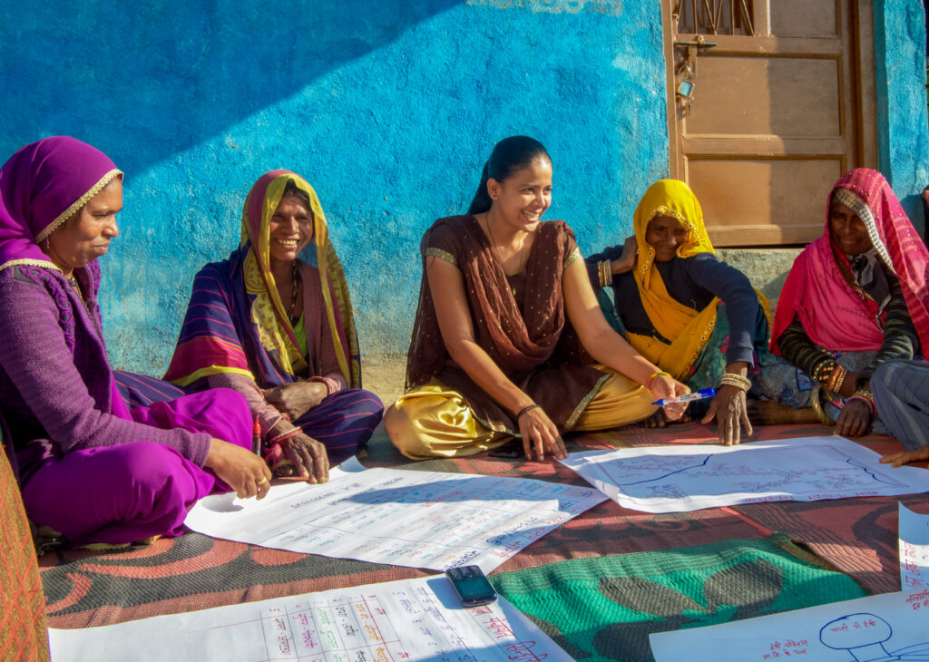 Five women sit on a mat and study several large documents that are laid out in front of them.